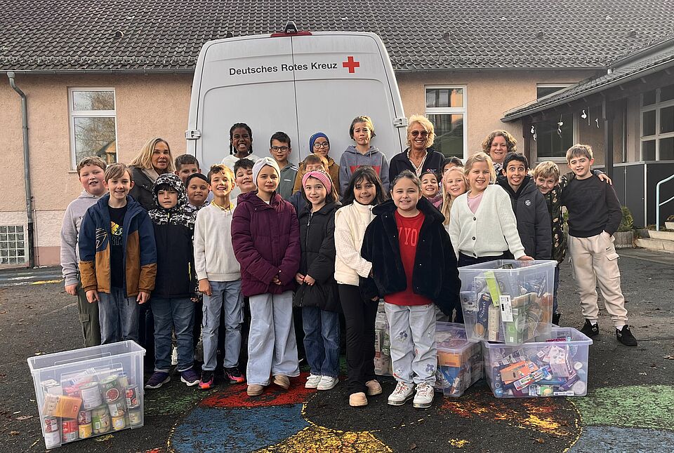 Tafelladen_Foto_cbs.jpg Kinder der Brentano-Schule stehen vor deinem Rotkreuzfahrzeug, daneben die gespendeten Waren in Kisten. Dabei sind die Schulleitung und Vertreter des DRK